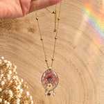 Può includere: Una collana color oro con una catenina sottile e piccole perline dorate distanziate uniformemente. Il ciondolo è un pezzo di resina trasparente di forma ovale contenente fiori pressati rosa e bianchi, decorato con due perle e una pietra.